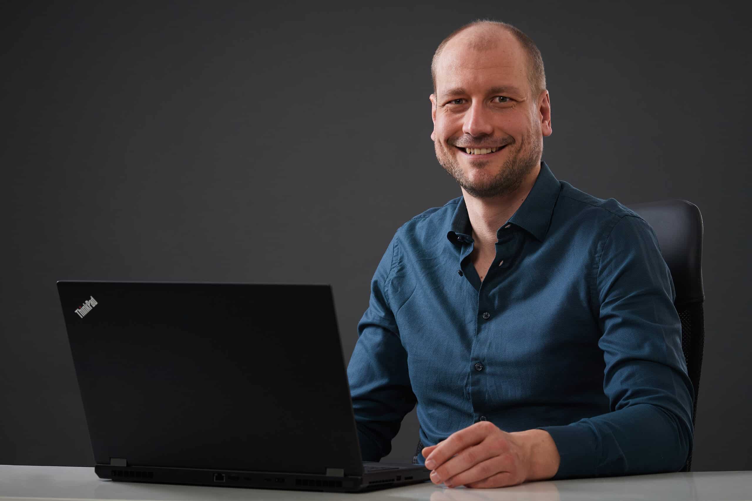 Portrait Christopher Schulz sitzend am Laptop vor dunklem Hintergrund